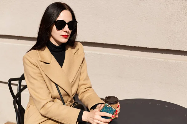 Bild av ung vacker brunett kvinna i svart solglasögon och beige snygg kappa sitter vid bordet i utomhus kafé, dricka hett säljare från Paper Cup, med middagspaus. Människor Concept. — Stockfoto