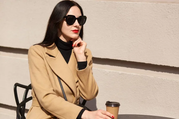 Foto al aire libre de la mujer morena con el pelo largo con abrigo beige, sentado en la mesa en la cafetería de la calle y la celebración de café para llevar, atractiva hembra tiene expresión facial pensativa, mantiene la mano bajo la barbilla . — Foto de Stock