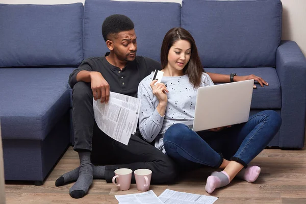 Interracial couple of young people sitting together in front laptop on floor near sofa, in their appartments, busy with composing financial report, using paper documents and portable computer, — Stock Photo, Image