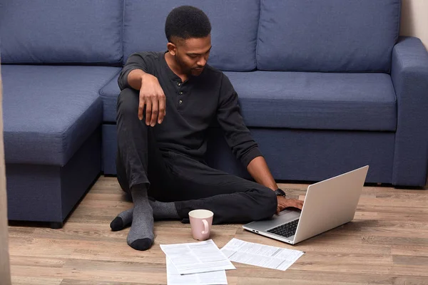 Horizontale foto van donkere gevild jonge knappe man doet papier thuis, controleren van documenten, drinken van warme drank, zittend in de buurt van grote blauwe Bank op houten vloer. Mensen en online werk concept. — Stockfoto