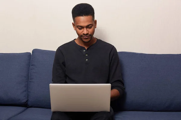 Indoor shot of dark skinned young male working online at home, surfing internet on portable laptop computer, sitting on comfortable blue couch, handsome man dressed casually. People and work concept.