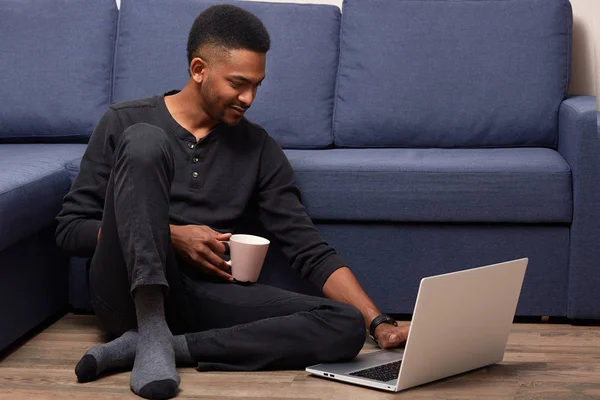 Donkere gevild jonge man werken met zijn laptop thuis, houdt beker met warme drank, zittend in de buurt van de Bank op de vloer, knappe man gekleed terloops, het controleren van zijn e-mail of het kiezen van film voor kijken. — Stockfoto