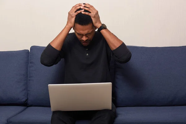 Indoor shot of stressed black young male, keeps hands on head, tries to concentrate, wearing black casual jumper, working on laptop computer, using wireless Internrt, sitting on comfortable sofa.