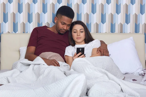 Young attractive multicultural couple in white bed, female holding smartphones in hands, wearing pajamas, searching or browsing Internet or watching film together, cheating over messages with friends.