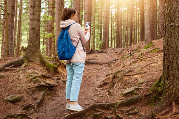 Obraz zkušeného cestovního blogera stojícího uprostřed lesa kolem obrovských stromů, který si přidržoval telefon v obou rukou, fotografoval pro společenské sítě a cestoval sám po cestě. Cestování — Stock fotografie