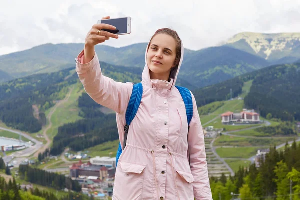 Zachwycony cute młoda kobieta z przyjemnym uśmiechem na twarzy stoi na szczycie wzgórza, trzymając jej smartphone, patrząc na aparat uważnie, robienia zdjęć na tle górskim. Koncepcja blogowania podróży. — Zdjęcie stockowe