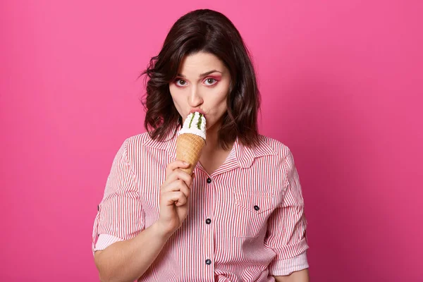 Retrato de morena concentrada seria sosteniendo helado, probándolo, mirando directamente a la cámara, teniendo maquillaje brillante, usando ropa casual, de pie aislado sobre fondo rosa en el estudio . — Foto de Stock