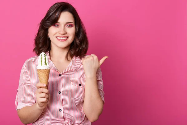 Estúdio interno tiro de positiva bela jovem de pé isolado sobre fundo rosa, segurando sorvete em uma mão, fazendo gesto com o polegar, sorrindo sinceramente. Espaço de cópia para propaganda . — Fotografia de Stock