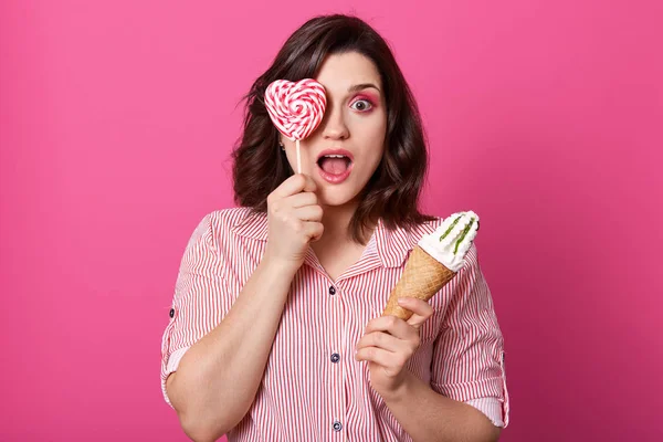 Divertido impresionado hermosa mujer de pie aislado sobre fondo rosa en el estudio, abriendo los ojos y la boca ampliamente con shock, cubriendo un ojo con piruleta en forma de corazón, la celebración de helado . — Foto de Stock