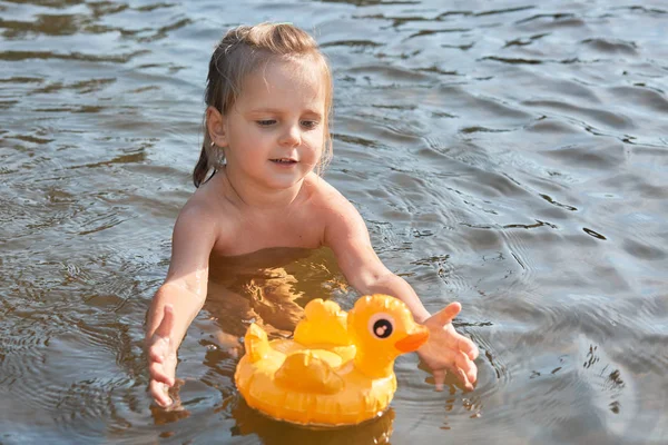 열정적 인 활기찬 작은 아이는 혼자 물에서 수영, 깨끗한 강에서 휴식을 즐기고, 자연과 일치하여 여름 휴가를 보내고, 장난감을 가지고 놀고, 물 표면에 고무 오리를 가지고 있습니다.. — 스톡 사진