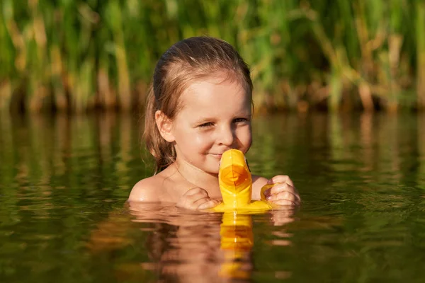 좋은 찾고 긍정적 인 아이 혼자 지역 호수에서 수영, 물에 대한 그녀의 고무 오리를 들고, 여름 휴가를 즐기고, 갈대 근처 수영, 높은 영혼에있는. 어린이 및 물 게임 개념. — 스톡 사진