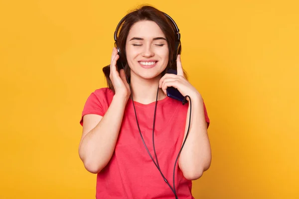 Souriante jeune femme joyeuse, isolée sur fond jaune en studio, fermant les yeux en écoutant de la musique, ayant des écouteurs, prenant du plaisir, portant un t-shirt rouge décontracté. Temps libre . — Photo