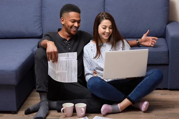 Indoor shot of young beautiful couple, sitting on floor, using their laptop, having pleasant communication, smiling sincerely, looking at laptop screen, spending weekends together, making plans. — Stock Photo, Image