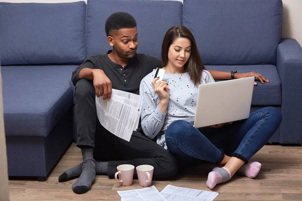 Young brunette bussinesswoman staying at home with her husband, having some work, applying credit card information, making purchases online, man sitting near his wife being serious and confused. — Stock Photo, Image