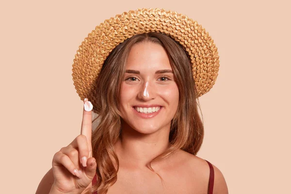 Young woman applaying sunblock on her face, and showinig her finger with sunscreen, model posing isolated over beige background, standing against studio wall with topothy smile, dressed swimming suit. Stock Photo