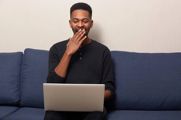 Young freelancer in casual clothes, sitting on blue sofa in front of laptop and yawning, looks tired and exhausted, having lots work to do. Handsome dark skinned female working online. people concept. Stock Photo