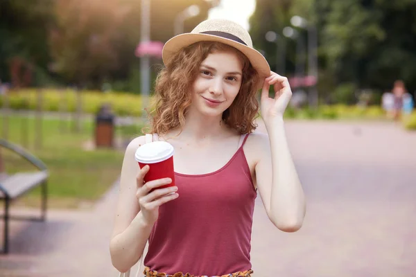 Portrait of magnetic positive female smiling sincerely, holding papercup of takeaway drink, having coffee break, touching her straw hat, spending time outside, looks peaceful and friendly. Break conce Stock Picture