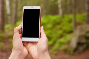 Akıllı telefonu tutan ellerin açık hava resmi kapatıldı, onu yükseltti, açmaya çalıştı, bağlantı bulmaya çalışıyor, yeni bir cihaz kullanıyor, boş ekrana sahip, doğa arka planı üzerinde mobil. Copyspace.
