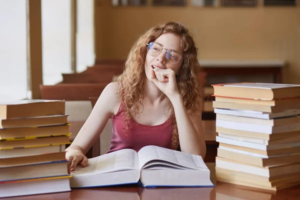 Portrait of thoughtful dreamy young student sitting in reading hall, putting her hand close to face, touching huge opened book, looking out of window, preparing for exam. Students life concept. — Stock Photo, Image