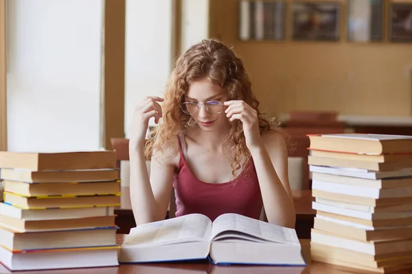 Close up portrait of young woman touching her eyeglasses, reading paper book. Female student spending long hours, wearing glasses, casual burgundy t shirt, preparing for test or exam in reding room. — Stock Photo, Image