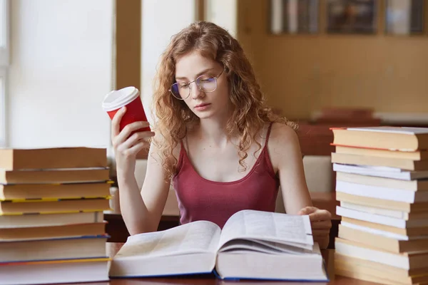 Jeune femme attirante portant des lunettes et uotfit décontracté, livre de lecture qui repose sur la table et tenant tasse de café dans sa main tout en étant assis près du bureau en bois avec pile de livres sur elle, près de la fenêtre . — Photo