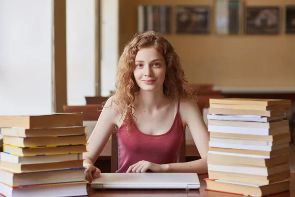 Prise de vue d'une jeune étudiante poilue assise à table à la bibliothèque, se préparant au test ou à l'examen, posant avec une expression faciale agréable, regardant diéremment concentrée à la caméra. Concept d'éducation . — Photo