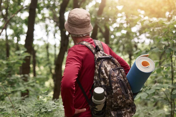 Foto esterna di vecchio che ha borsa con thermos e piumino, indossa cappello beige e felpa rossa, in cerca di avventure nella foresta da solo, essendo appassionato di viaggi ed escursioni. Concetto di hobby . — Foto Stock