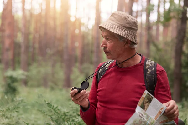 Escursionista che indossa maglione casual rosso e cappello, posa con lo zaino, cerca la giusta direzione con la bussola, perdendosi nella foresta, illuminato dalla luce dorata del sole. Stile di vita attivo e concetto di vita moderna . — Foto Stock