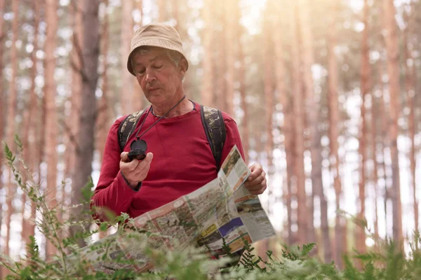 老人徒步旅行和看指南针和地图的方向在森林旅行的图像，高龄男性穿着休闲装与背包丢失在木材。旅游和积极的娱乐理念. — 图库照片