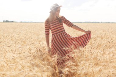 Şık çizgili elbise ve saman şapka güneşli yaz gününde buğday alanında yürüyüş genç güzel kız Görüntü, gün batımı zevk, model çayır açık poz, kenara bakıyor. Özgürlük kavramı.