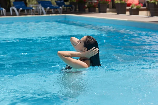 Perfil de mujer joven y elegante de pelo negro nadando en la piscina, pasando tiempo en el lujoso hotel spa resort, tocándose el pelo con ambas manos, tomando el sol en el agua. Concepto de tiempo de vacaciones . — Foto de Stock