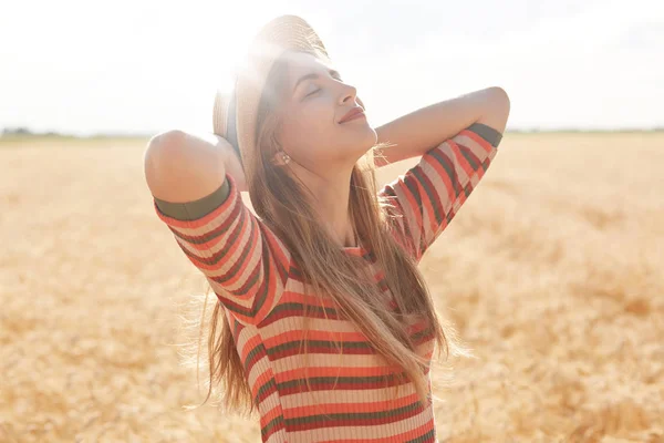 Tiro ao ar livre de mulher jovem feliz em roupa listrada e chapéu de sol desfrutando do sol no campo de cereais, fêmea posando com as mãos para cima, olhando sonhadoramente para o céu. Natureza, verão, férias e conceito de pessoas . — Fotografia de Stock