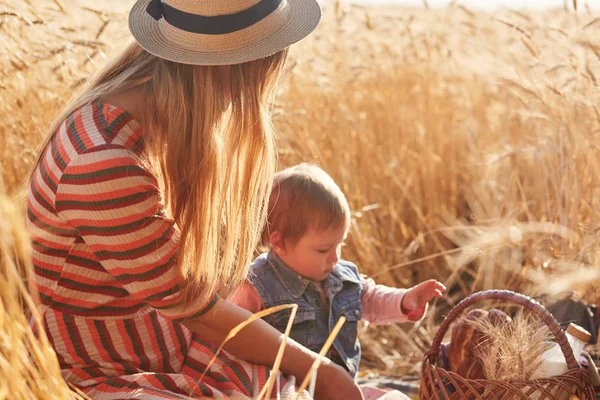 Outdoor picture of young fair haired mother sitting with her little daughter at wheat field, having small picnic together, mum looking after kid, going to eat and drink outside, spending good time. — Stock Photo, Image
