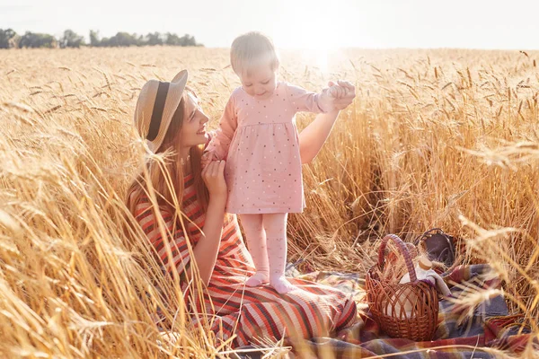 Happy mother looking at her daughter, holding her with hands, smiling sincerely, enjoying family rest, spending time with pleasure, taking basket with products, playing on blanket. Family concept. — Stock Photo, Image