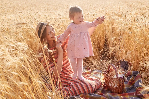 Outdoor shot of beautiful woman with her little doughter toddler wearing pink dress and tights in wheat field on summer day, mommy dressed striped dress and straw hat posing surrounded with spikelets. — Stock Photo, Image