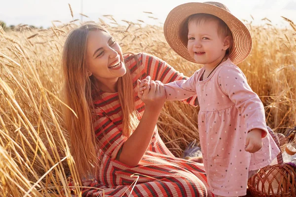 Picture of cute little child wearing pink smart dress, straw hat, smiling sincerely, spending time with her mother. Sweet cute mom looking at her daughter with love, laughing, holding her with hand.