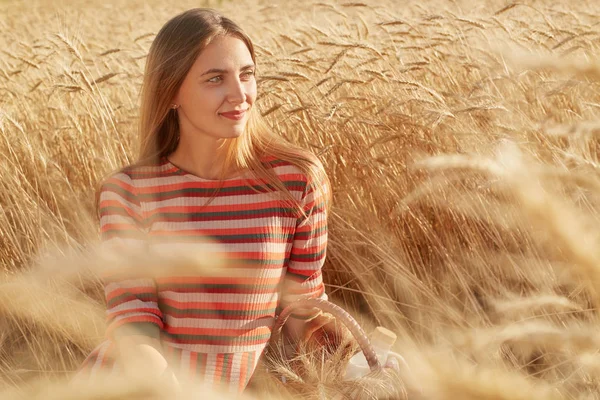 Retrato ao ar livre de mulher bonita adorável sentado no chão no campo de trigo, olhando para o lado, tendo expressão facial agradável, vestindo vestido colorido casual, apreciando a natureza ao redor. Conceito de natureza . — Fotografia de Stock