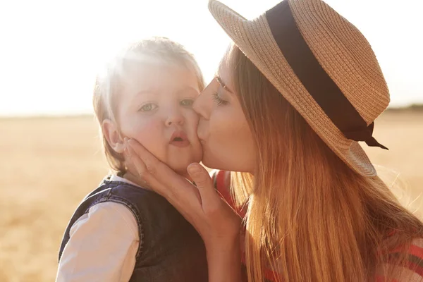 Outdoor picture of loving caring woman with straw hat on head kissing her little daughter, touching her face with hands, posing over wheat field background. Little kid looking directly at camera. — Stock Photo, Image