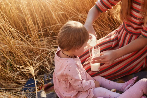 Portrait of cute fair haired kid looking attentively at her mother, waiting for having drink, caring mum filling glass with milk from bottle for child, having rest at wheat field sitting on blanket. — Stock Photo, Image