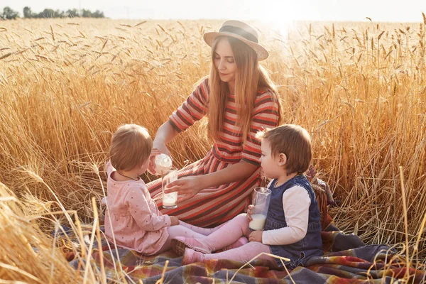 Caring helpful mother sitting on blanket at wheat field with her lottle daughters, filling glasses with milk from bottle, wearing dress and hat, childen behaving in proper way. Motherhood concept. — Stock Photo, Image