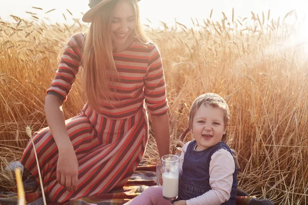 Image of happy cheerful infant looking directly at camera, holding glass of milk in one hand, being happy. Smiling good looking woman looking after her daughter, wearing stripped dress and straw hat. — Stock Photo, Image