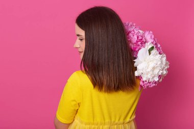 Parlak sarı elbise içinde poz veren ince genç Avrupalı modelin arka görünümü, bir kenara bakarak, omuzda beyaz ve pembe şakayık tutan, kısa siyah saçlı, stüdyoda pembe arka plan üzerinde izole.