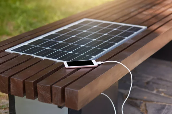 Outdoor shot of wooden bench in park having solar power panel installed, USB cabel connected to smartphone, modern device lying near solar panel charging, being switched off, having black screen.