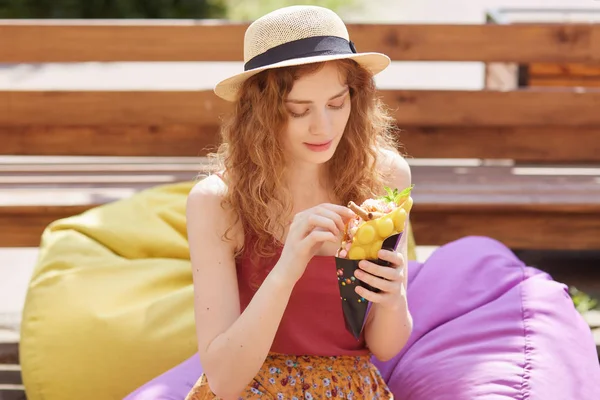 Bild av söt söt kvinna med Fair lockigt hår, tittar uppmärksamt på glass, med måltid, njuter av hennes helger ensam i parken, sitter på färgglada Beanbag, klädd i halm hatt. Ungdoms koncept. — Stockfoto