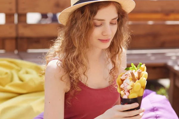 Outdoor shot of romantic cute young girl looking at her dessert, being in high spirits, feeling hunger, holding ice cream in hand, hanging out alone at cafe, posing over wooden fence background.