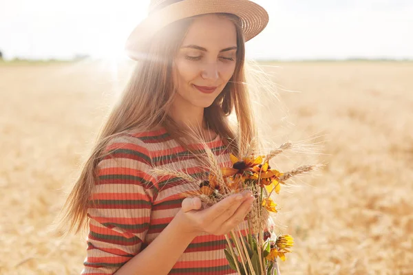 Pozitivní spokojené mladé ženy, oblečené do holených šatů a slamáku, pózující v pšeničném terénu, držící kytici, pečlivě se na ni, vychutnávají letní slunný den. — Stock fotografie