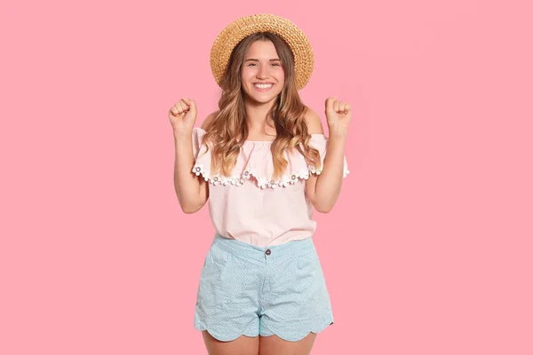 Studio girato di allegra femmina affascinante indossa cappello di paglia, camicetta leggera e pantaloncini, alzando le mani, essendo di buon umore, guardando direttamente la fotocamera, avendo i capelli ondulati. Concetto di persone ed emozioni . — Foto Stock