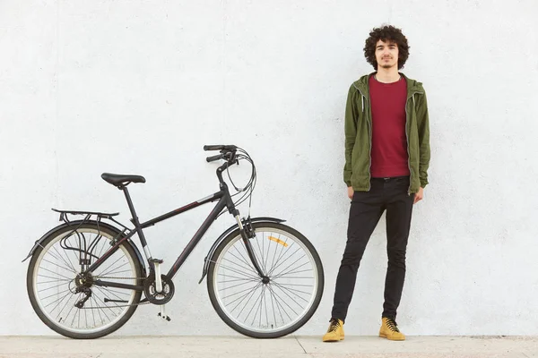 stock image Image of curly haired calm young boy standing near his bicycle, wearing black trousers, brown boots, red sweatshirt and green jacket, posing isolated over white wall outdoors. Sport concept.