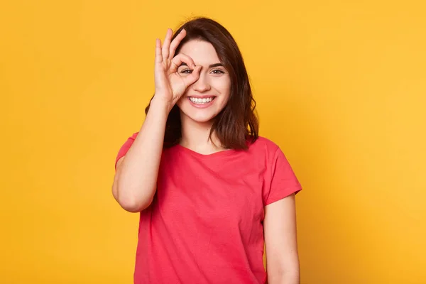 Imagen de una joven modelo alegre y tranquila posando aislada sobre un fondo amarillo brillante en el estudio, poniendo sus dedos a la cara, haciendo un gesto bien, sonriendo sinceramente, usando una camiseta casual roja . — Foto de Stock