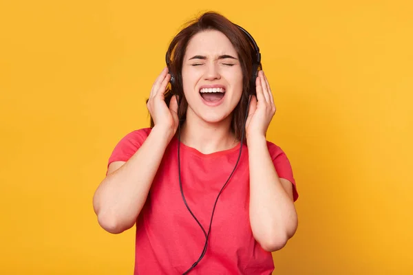 Imagen de atractiva morena enérgica con camiseta roja casual, con auriculares, escuchando música, cantando, disfrutando pasar su tiempo libre sola, cerrando los ojos, abriendo la boca ampliamente . — Foto de Stock